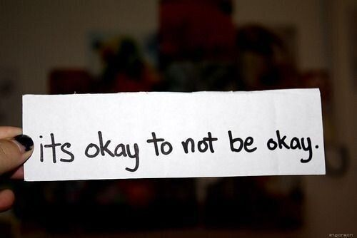 A hand holds a white piece of paper with the handwritten message 'it's okay to not be okay.' The background is slightly blurred, creating a soft and empathetic feel